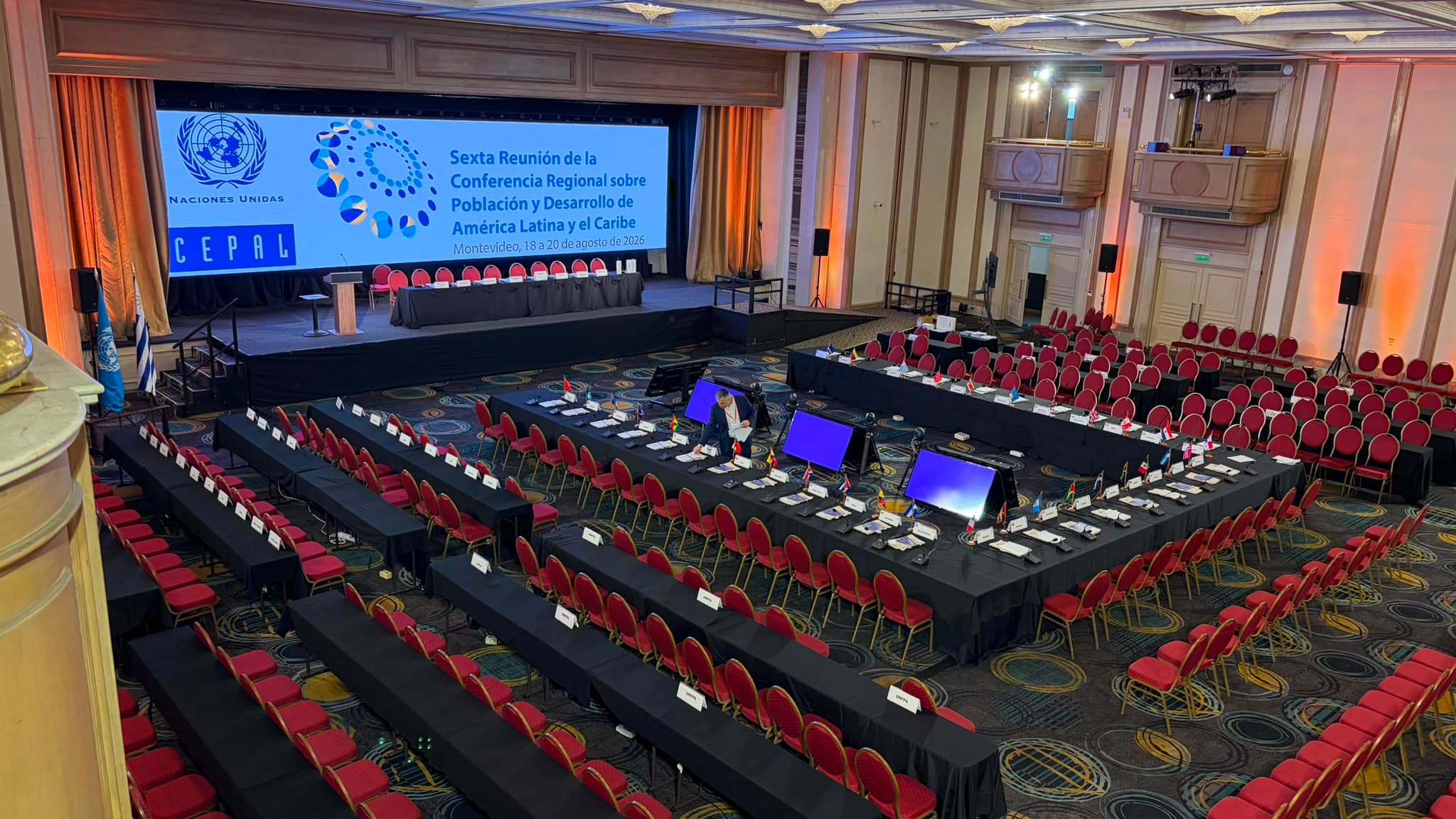 Salón montado para una conferencia internacional: pantalla LED de fondo y mesas en U con banderas y micrófonos de conferencia