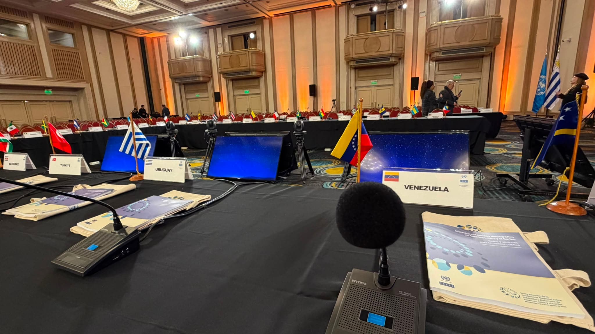 Detalle de la mesa de delegaciones, con micrófonos de conferencia, carteles de país y monitores de retorno