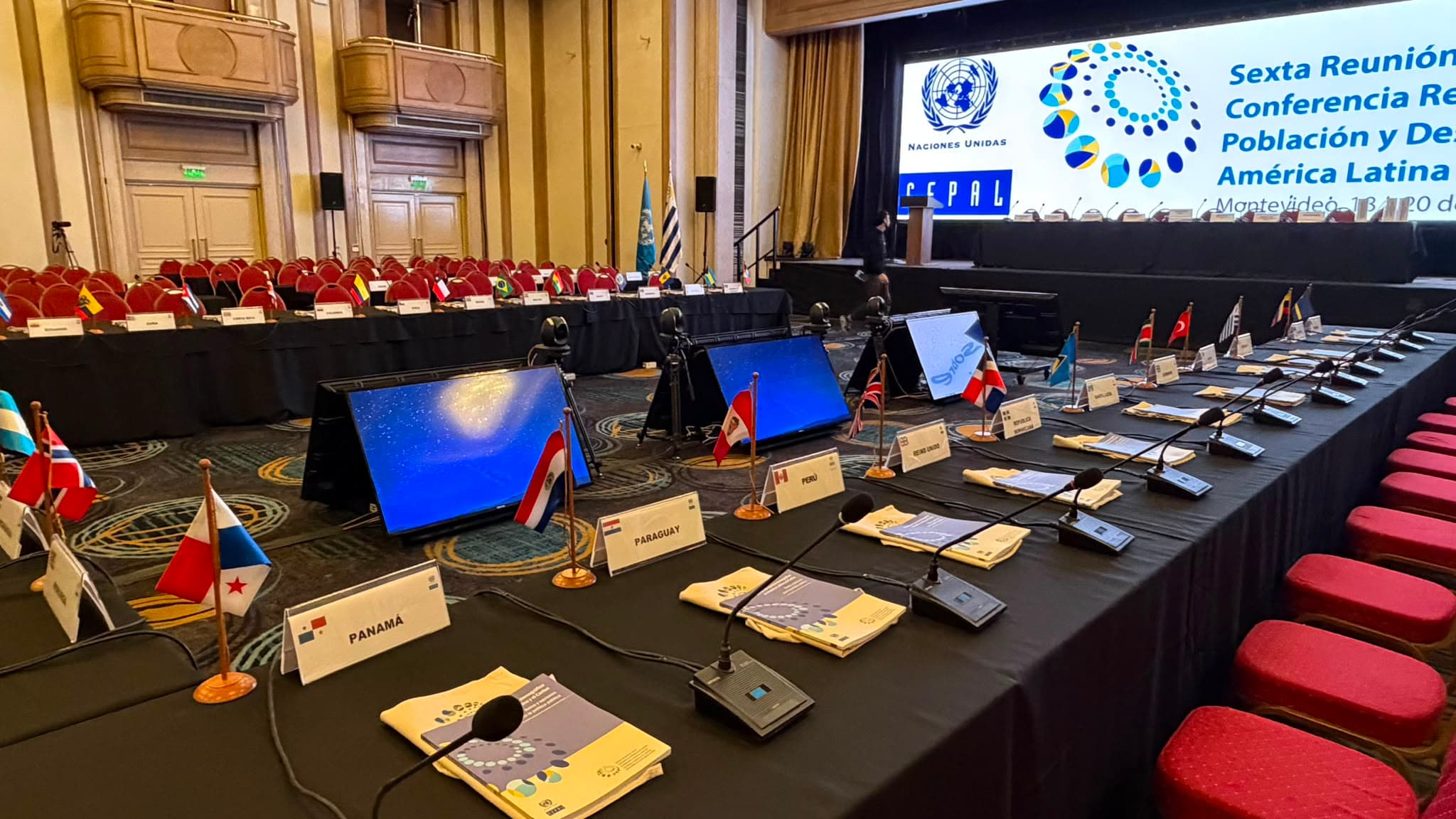 Mesa de delegaciones vista a lo largo, con banderas, micrófonos y la pantalla del escenario al fondo